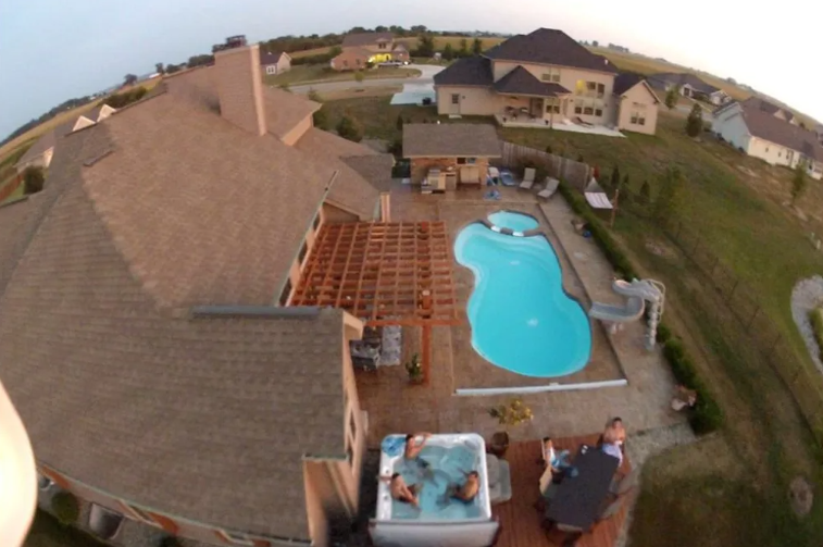 Aerial view of a backyard with a pool, hot tub, and pergola. People are in the hot tub.