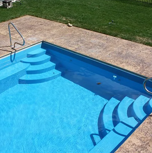 Blue rectangular swimming pool with steps, handrails, and surrounding concrete patio; grass in background.