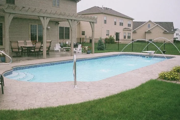 Backyard pool with fountain jets, patio, and houses in background.