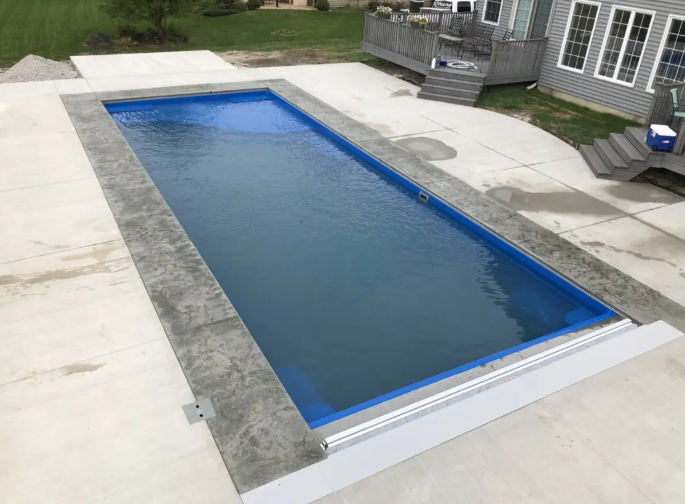 Rectangular blue pool surrounded by concrete, near a house with a deck.