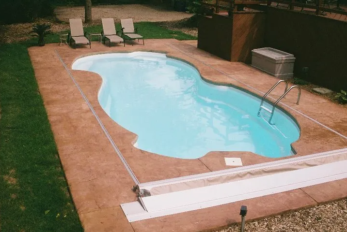 Swimming pool with a light blue interior, surrounded by brown concrete, lawn, and lounge chairs.