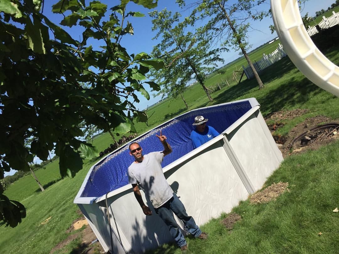 Two men inside a partially built blue-lined pool, waving. Outdoors in a grassy area with blue sky.