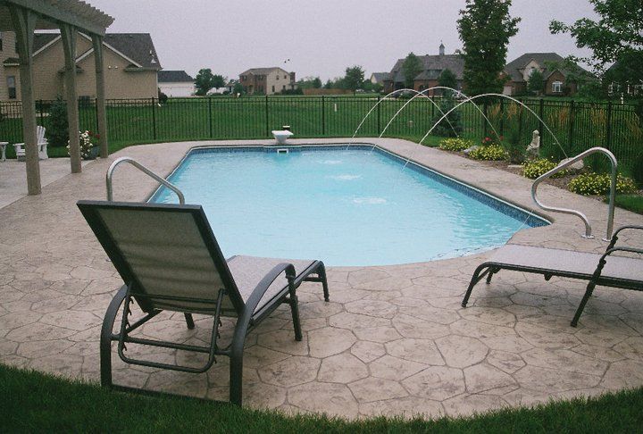 Rectangular swimming pool surrounded by stamped concrete patio, two lounge chairs, and water fountains.