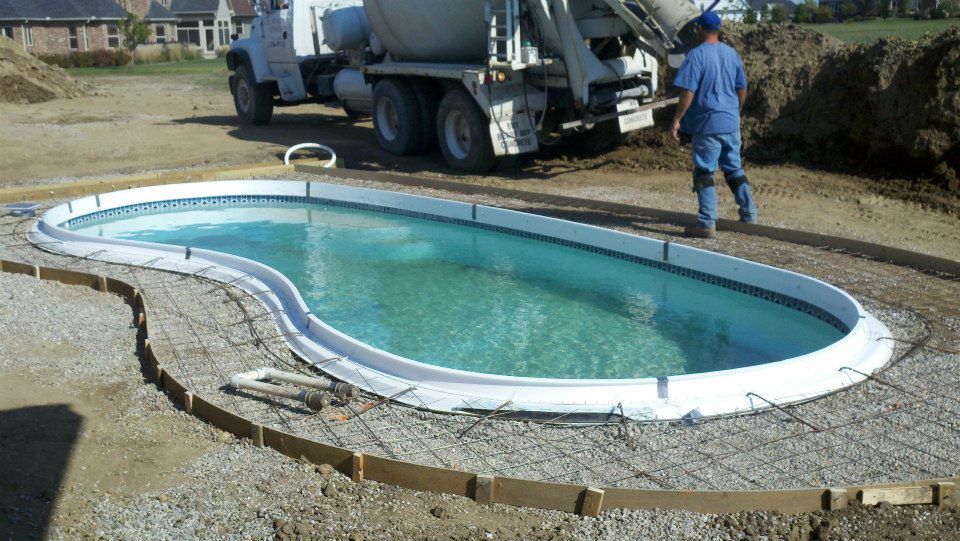 Concrete being poured into a swimming pool shell by a worker and a truck at a construction site.
