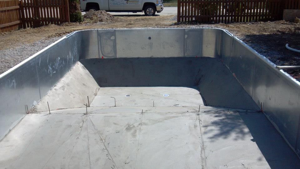 Empty rectangular pool under construction, metal and concrete, with surrounding yard and fence.