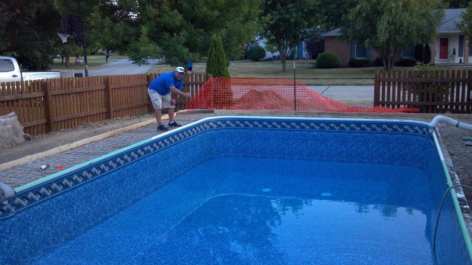 Man adding chemical to a blue, in-ground pool. Brown fence, orange safety net, and houses in the background.