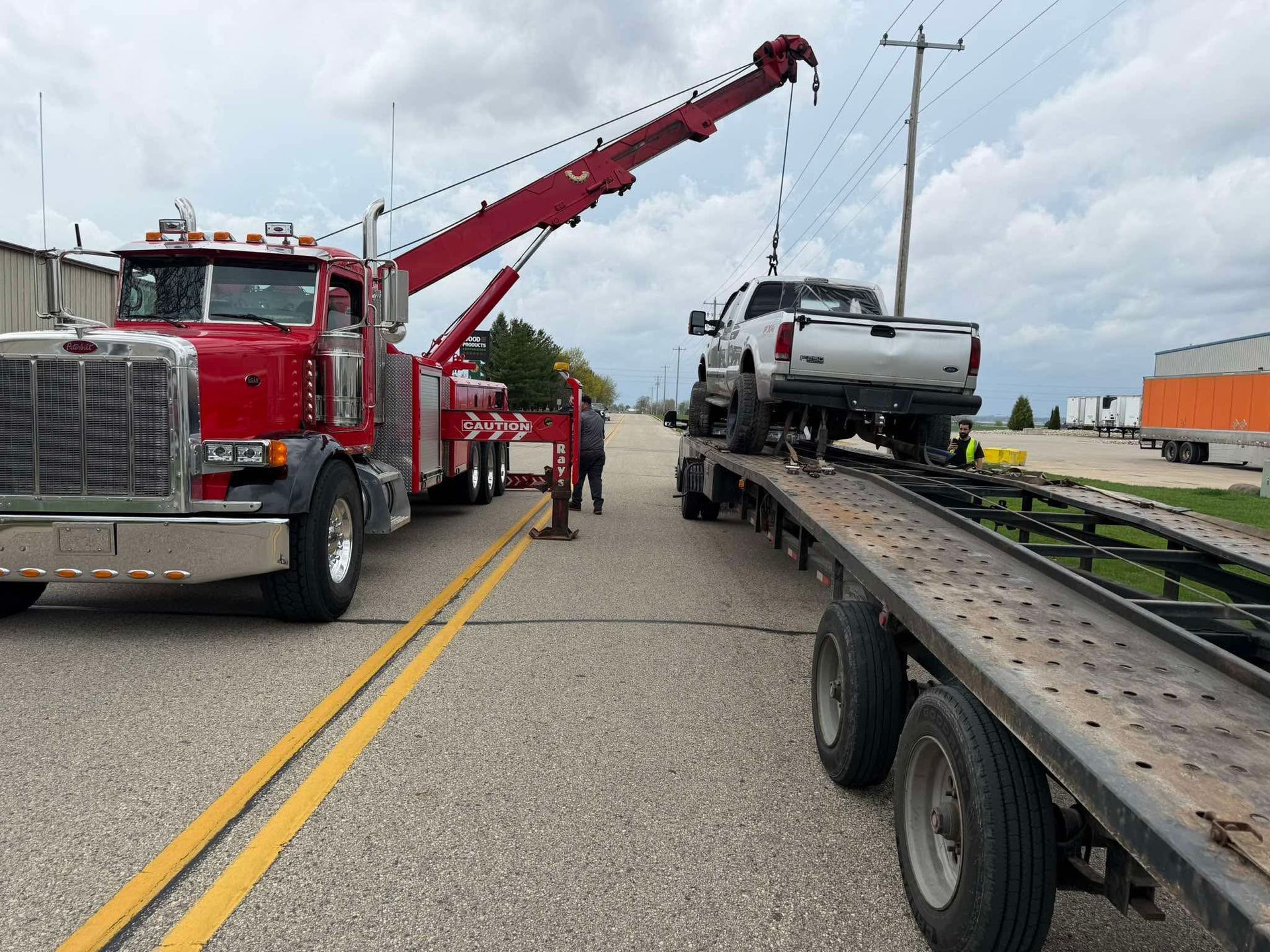 Tow truck lifting a silver pickup from a flatbed trailer on a road with workers nearby