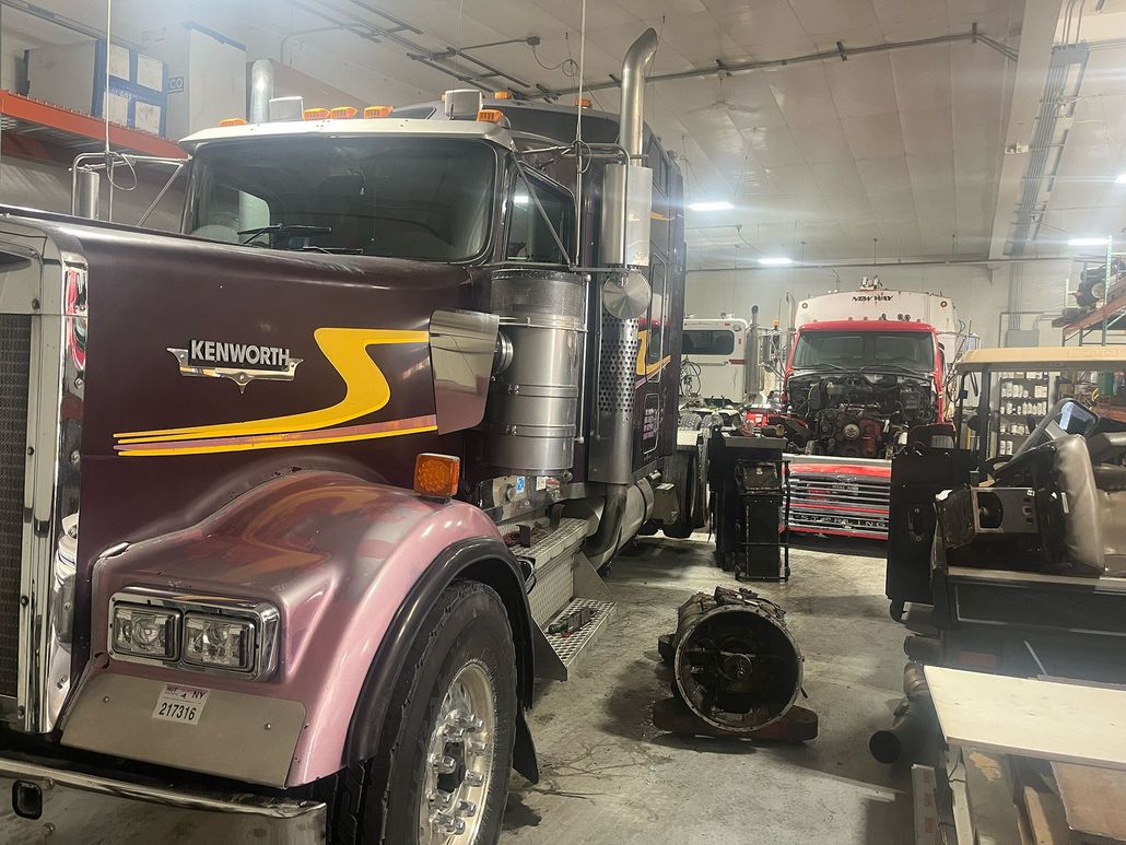 Semi-truck being repaired in a garage; a transmission sits on the floor.