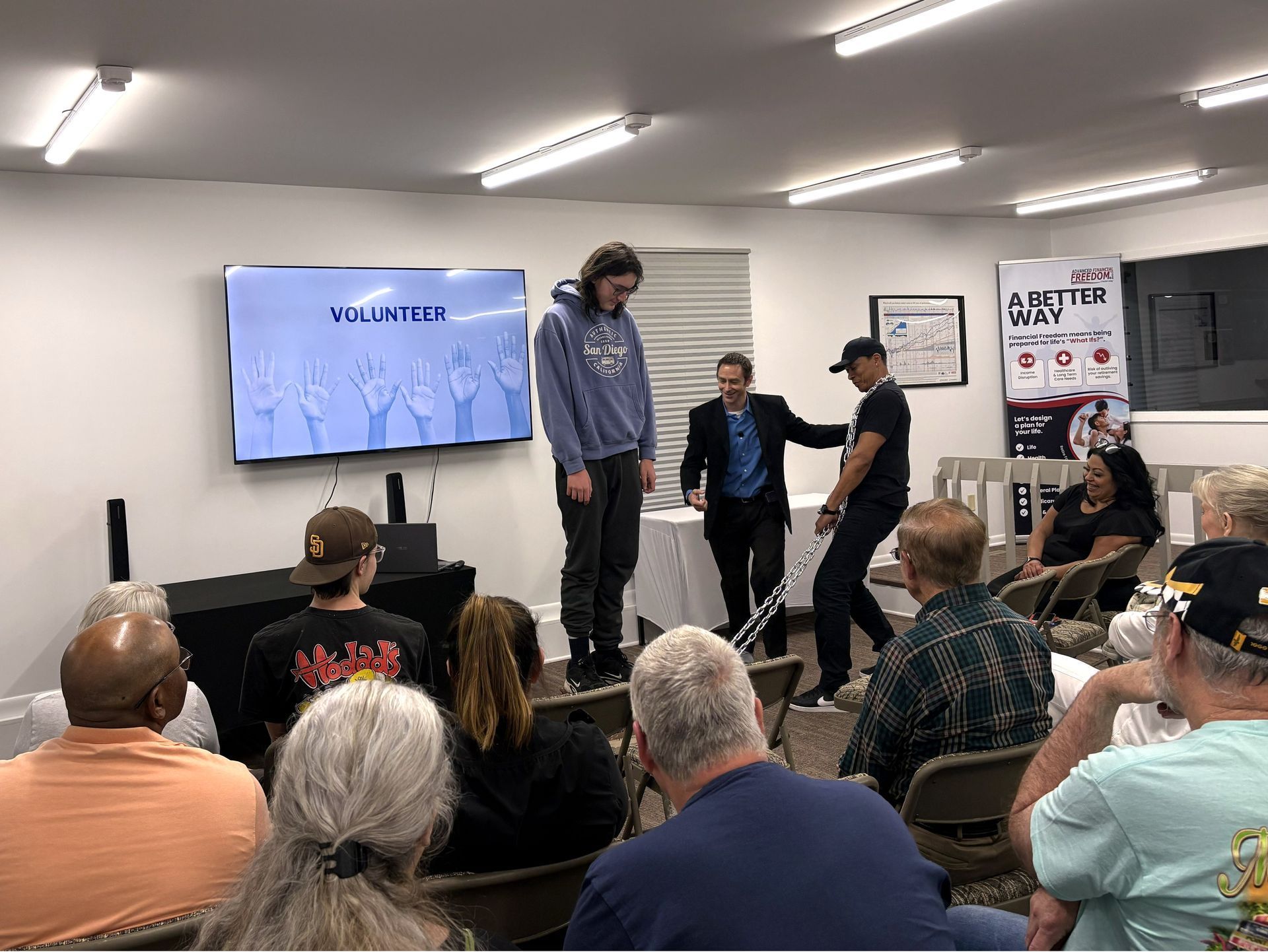 Audience watches two presenters on a small stage beside a wall-mounted screen in a meeting room.