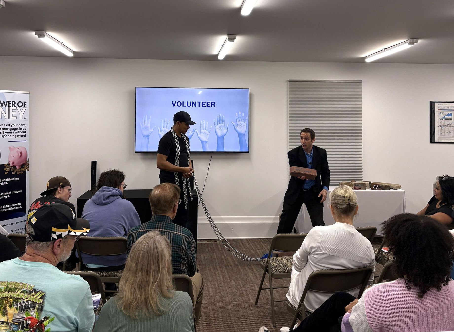 Two speakers on a small stage in a room, with an audience seated facing them and a screen behind them.