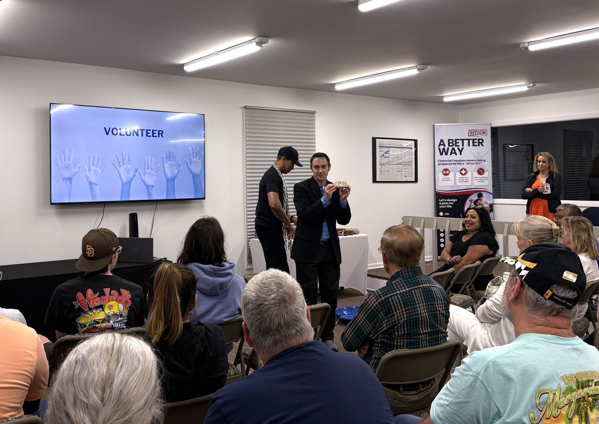 Audience watches two speakers presenting in a conference room with a projected slide and poster displays.