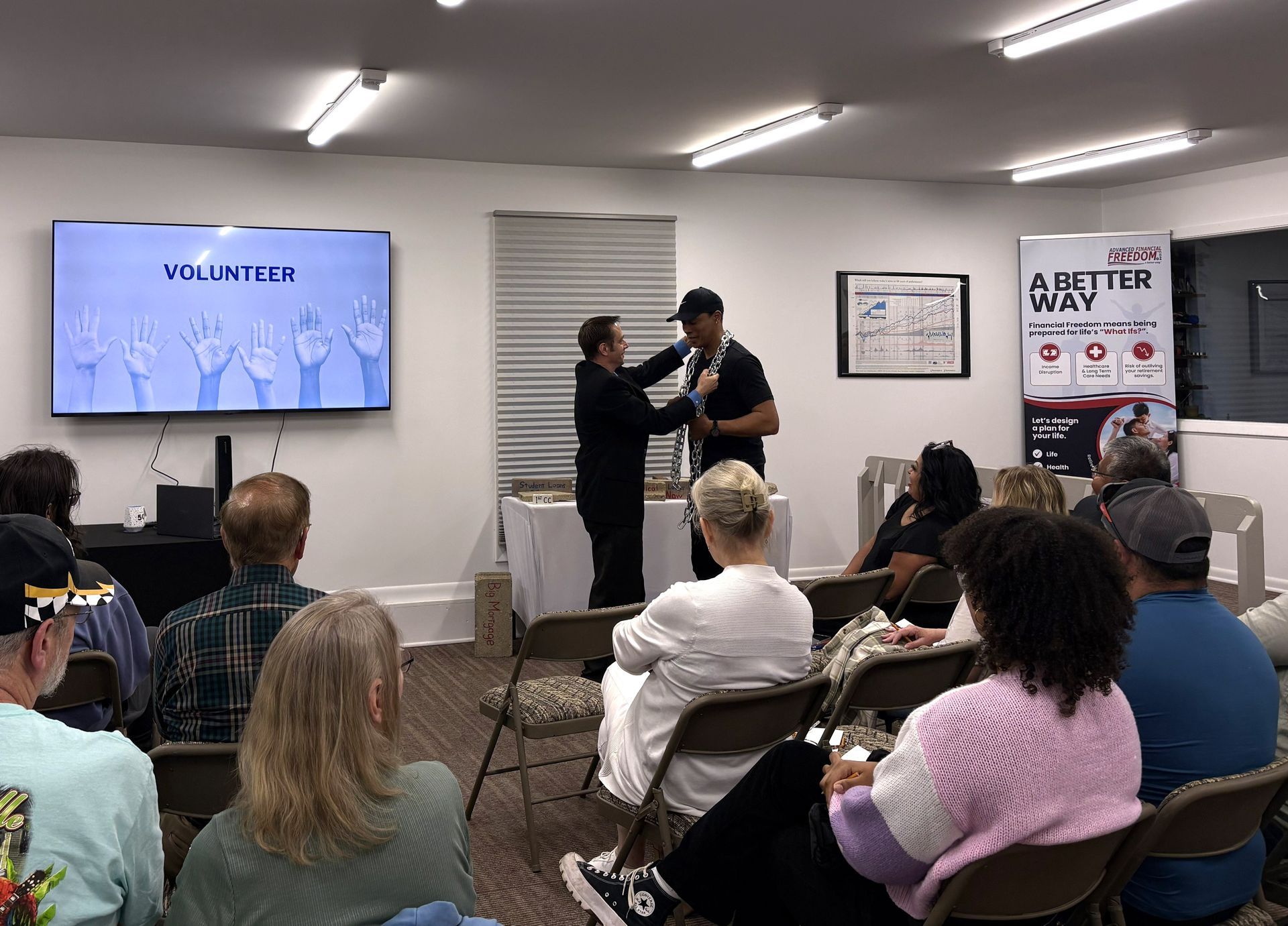 Audience at a small indoor event watches a speaker receive an item beside a screen and poster display.