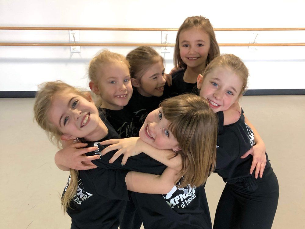 Six young girls in matching black shirts and pants, hugging and smiling in a dance studio.