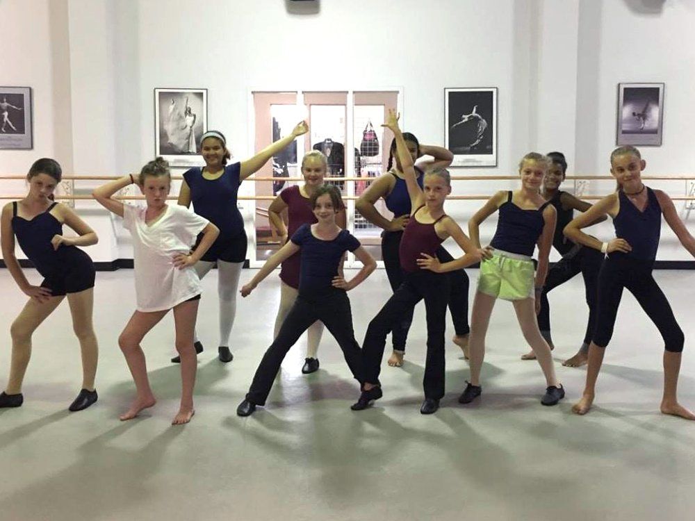 Group of young dancers posing in a studio. They wear various dance attire and have various expressions. Studio has mirrors and ballet pictures.