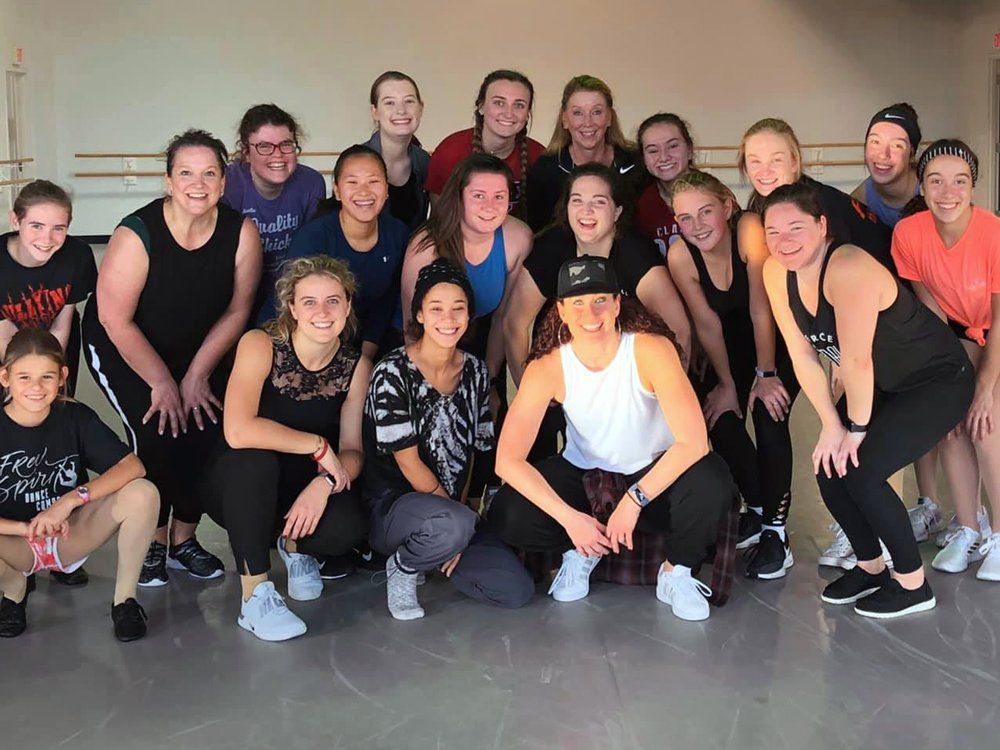 A group of young dancers, mostly female, pose together in a dance studio. They smile and wear athletic clothing.