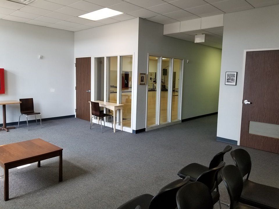 An empty, light-filled room with dark brown doors and a glass-walled space. Chairs and a table are in the foreground.