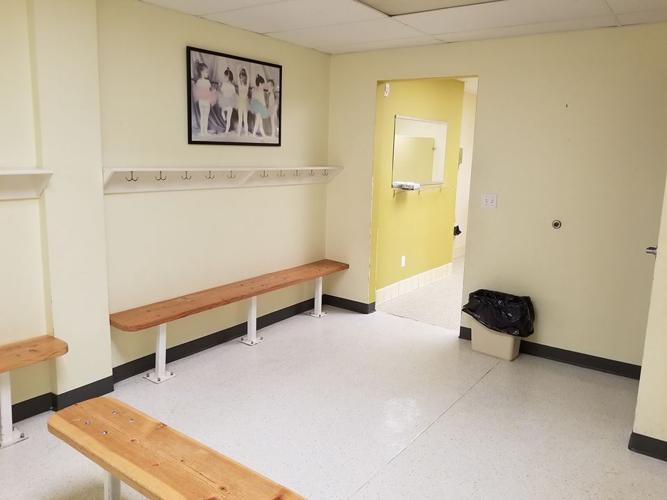 A yellow-walled changing room with wooden benches, a picture on the wall, and an open doorway to a yellow-walled space.