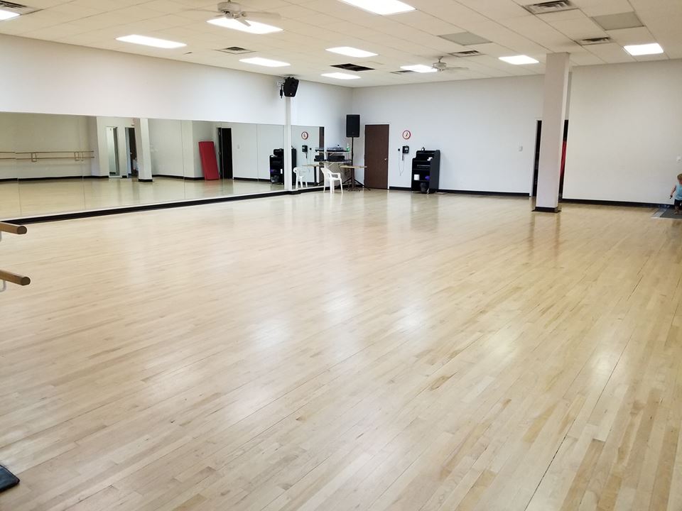 Empty dance studio with hardwood floors, mirrors, and ballet bar. White walls, several ceiling lights, speakers, and a doorway.