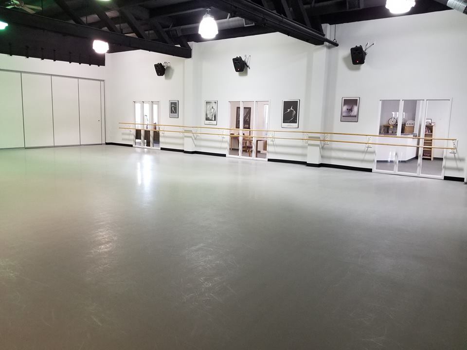 Empty dance studio with gray floor, white walls, and ballet barres along the walls.