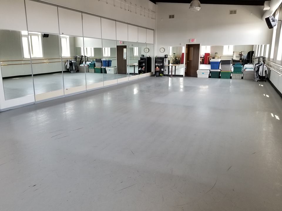 Empty dance studio with mirrors on one wall, gray floor, and windows along the other.