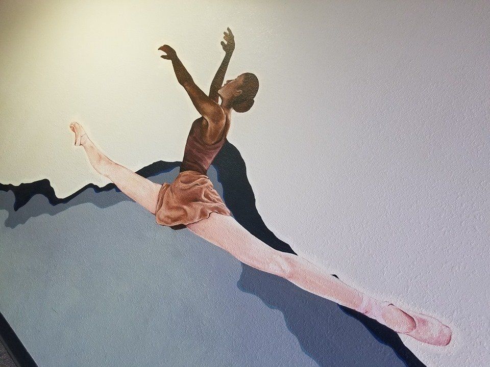 Ballet dancer mid-air in a split. She wears a pink leotard and skirt with arms raised. The background is a wall with shadows.