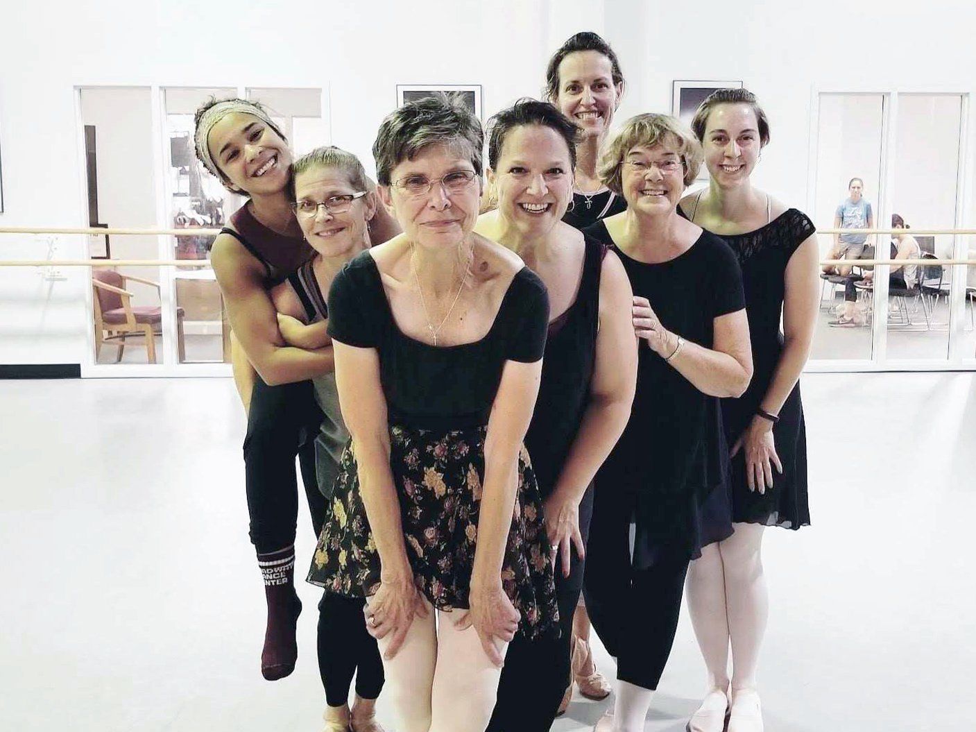 Group of eight women smiling in a dance studio; some are wearing leotards and skirts, ready for ballet.