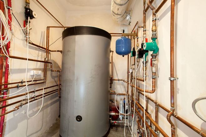 A tall, gray industrial water heater surrounded by a network of copper pipes and a blue expansion tank in a utility room.