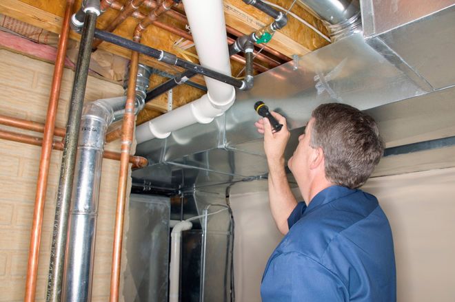 A worker in a blue uniform uses a flashlight to inspect pipes and metal ductwork on a basement ceiling.