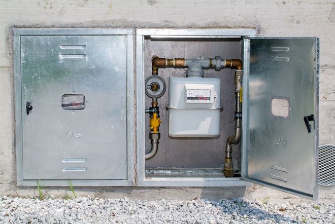 A gas meter encased in an open, dual-compartment metal utility box mounted on a concrete wall above gravel ground.