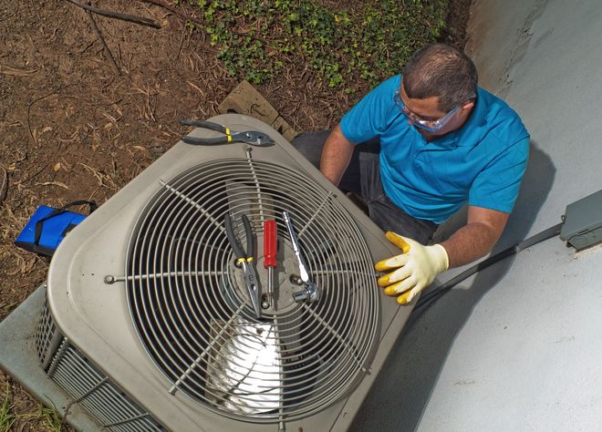 A technician in a blue shirt and protective gloves works on an outdoor air conditioning unit with various tools nearby.