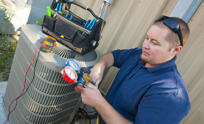 A technician in a blue shirt uses an HVAC gauge set to service an outdoor air conditioning unit next to a tool bag.
