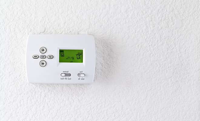 A white digital wall-mounted thermostat with a green display screen, mounted on a white textured wall.