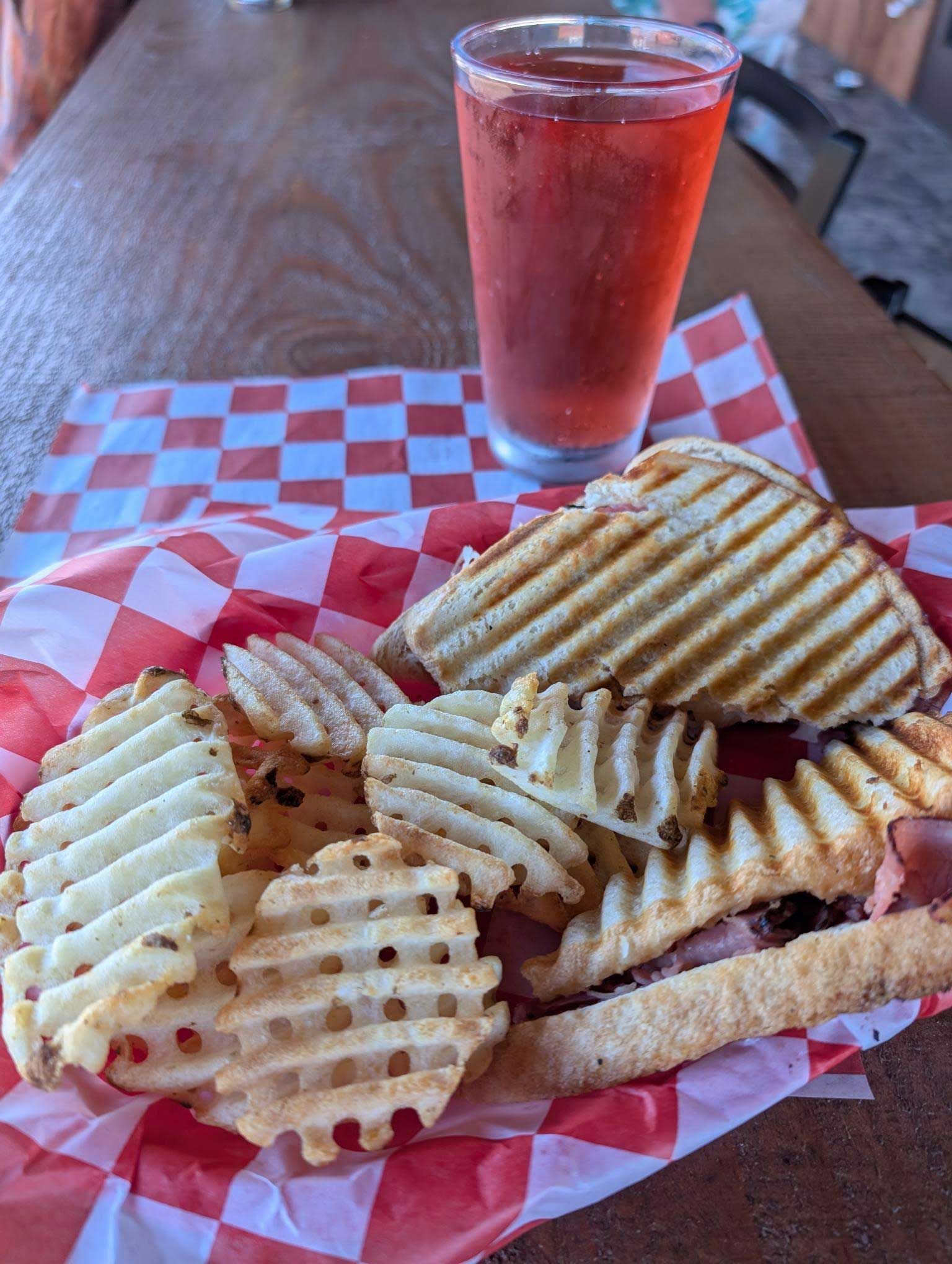 A panini sandwich and waffle fries on a checkered paper next to a glass of soda.