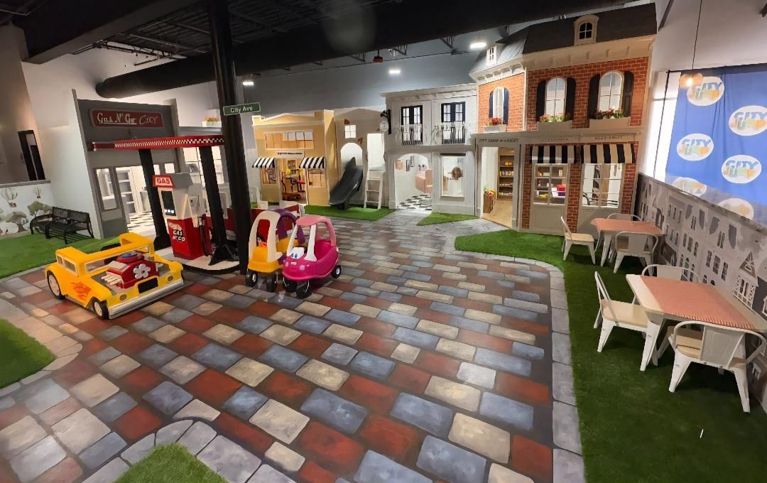 Indoor play area resembling a town square with toy cars, storefronts, and tables.