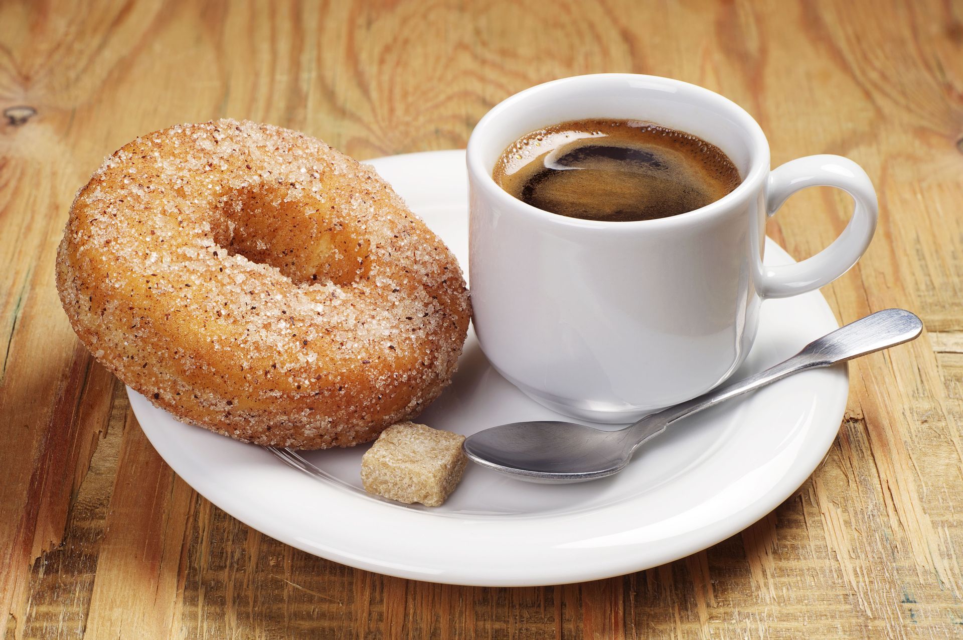 Donut and coffee on a white saucer with a spoon and sugar cube on a wooden table