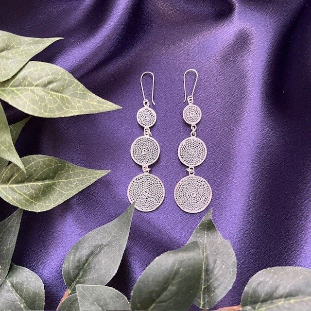 A pair of silver earrings are sitting on a purple cloth surrounded by green leaves.
