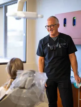 A dentist in black scrubs smiles, leaning near a patient in a dental chair. Office setting.