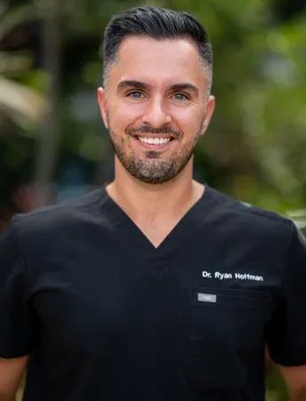 A man with a beard is wearing a black scrub top and smiling for the camera.