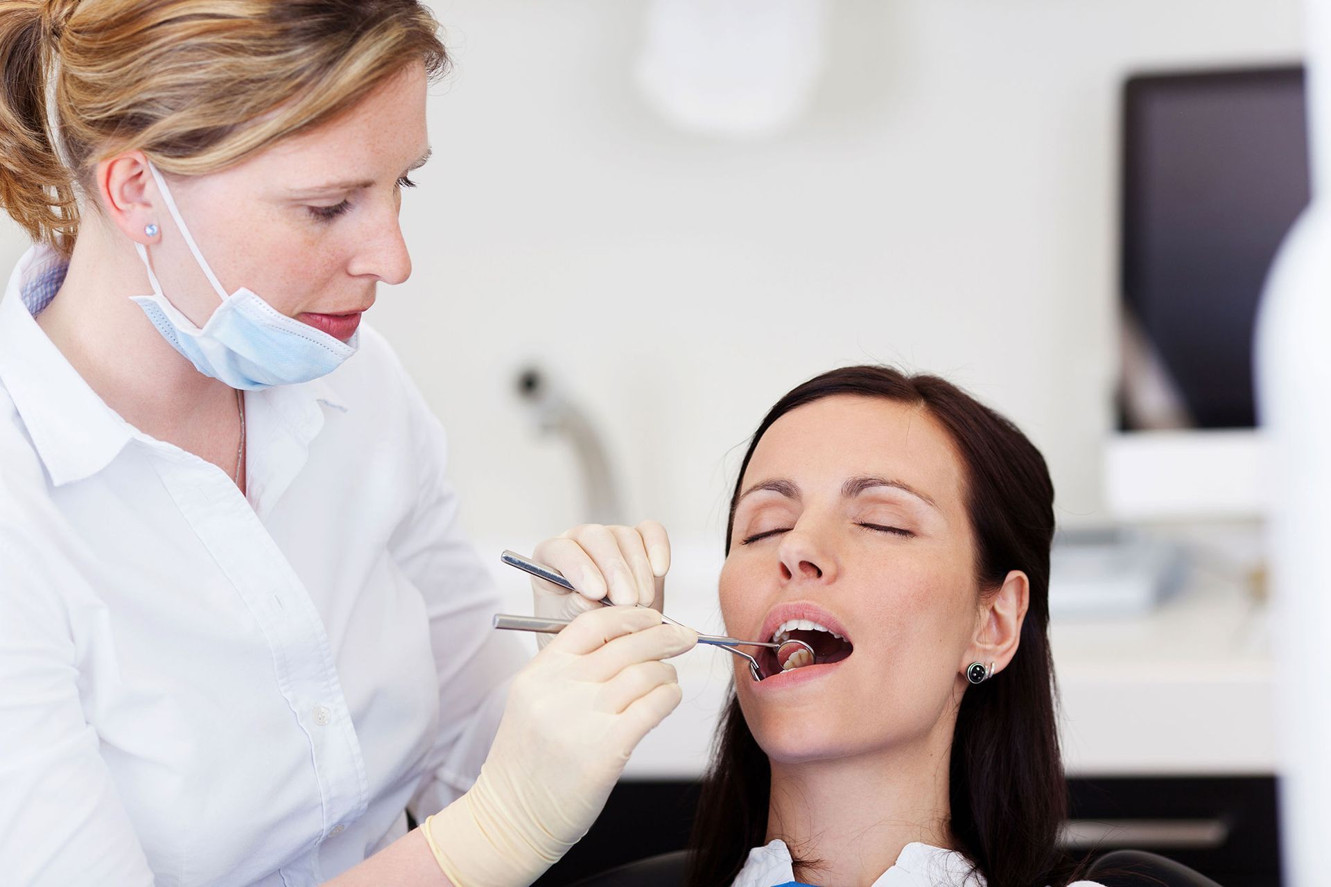 Dentist examining a woman's teeth in a dental office. The woman has her mouth open. The dentist wears a mask.
