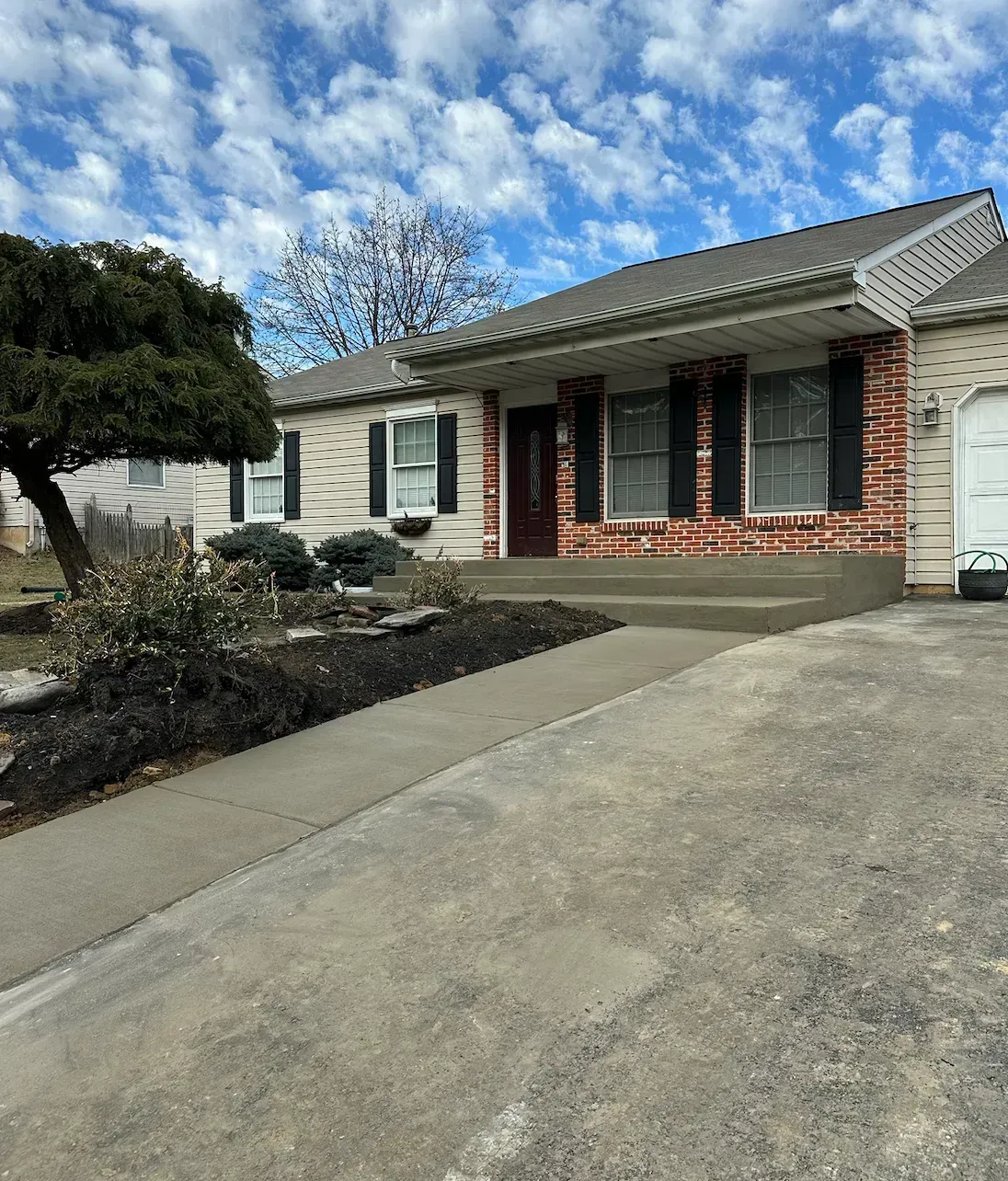 A single-story house with a brick facade and black shutters, a concrete walkway, and a cloudy blue sky.