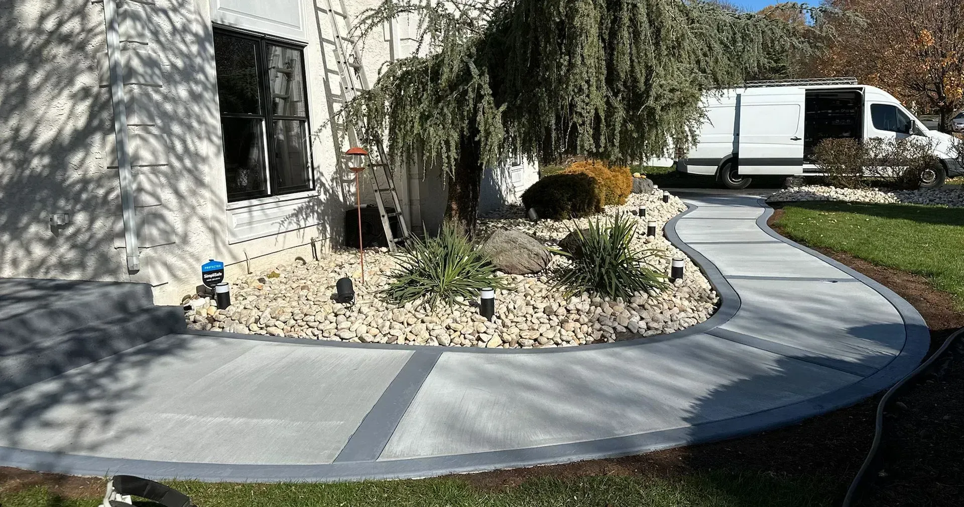 Curved concrete walkway with gray border, garden bed with light stones, weeping tree, and white van in background.