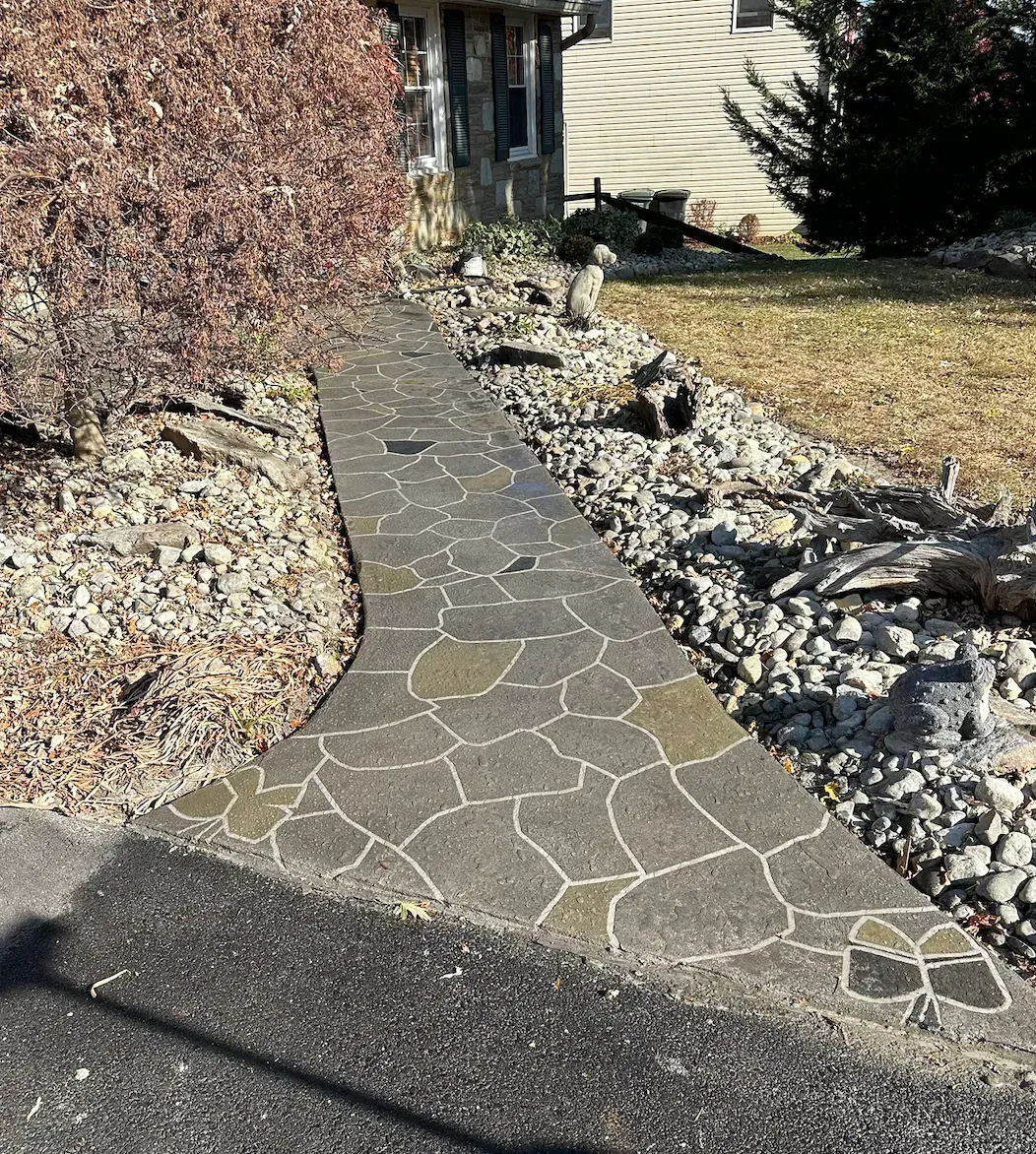 A paved walkway with a stone-like pattern, bordered by gravel and a house with beige siding.