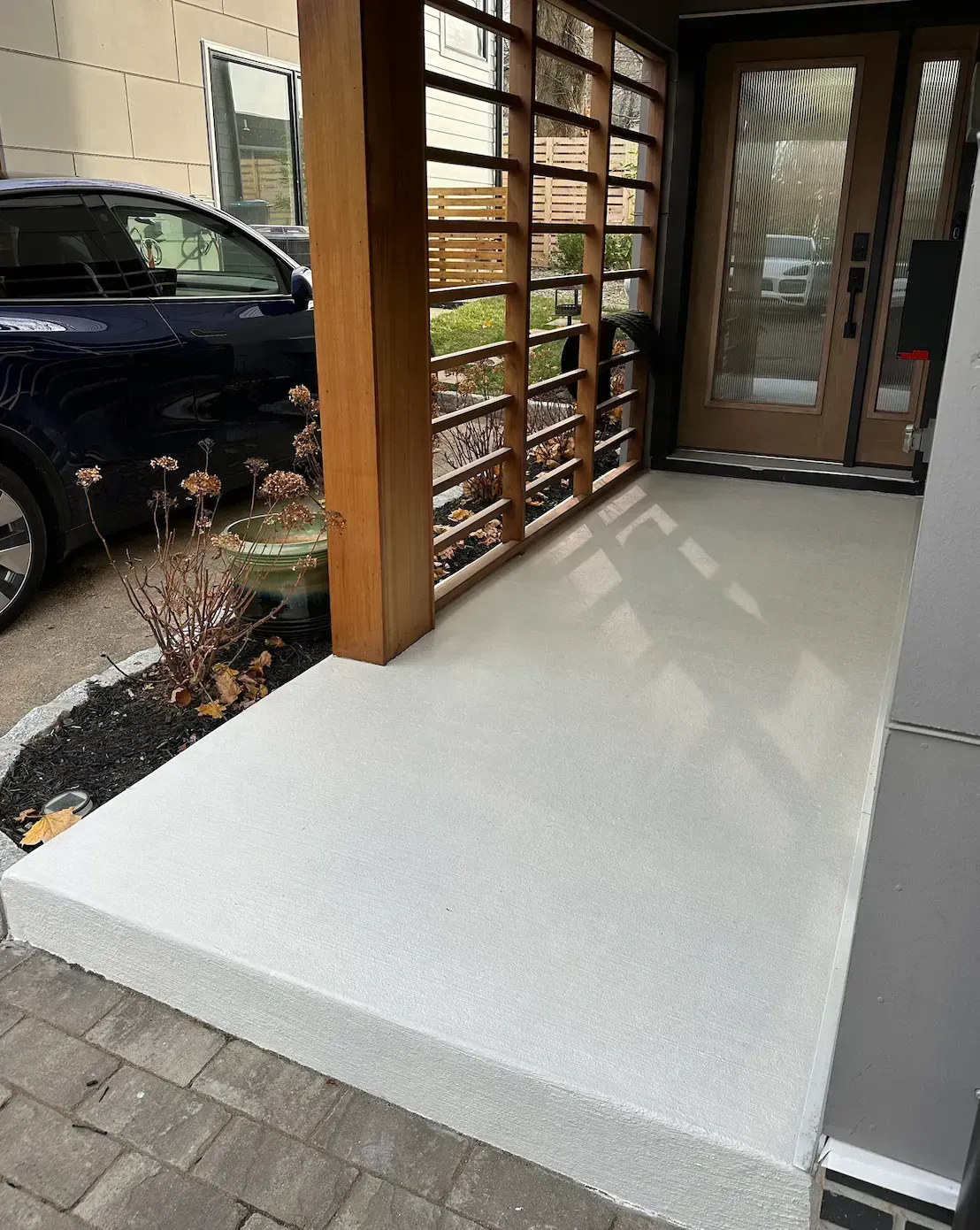 A light gray concrete porch with a wooden screen and a car parked to the left.