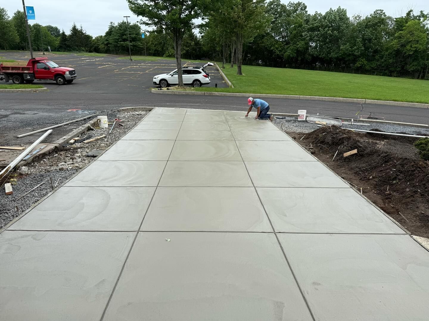 Concrete driveway under construction; worker kneels, smoothing surface; red truck, white car parked nearby.