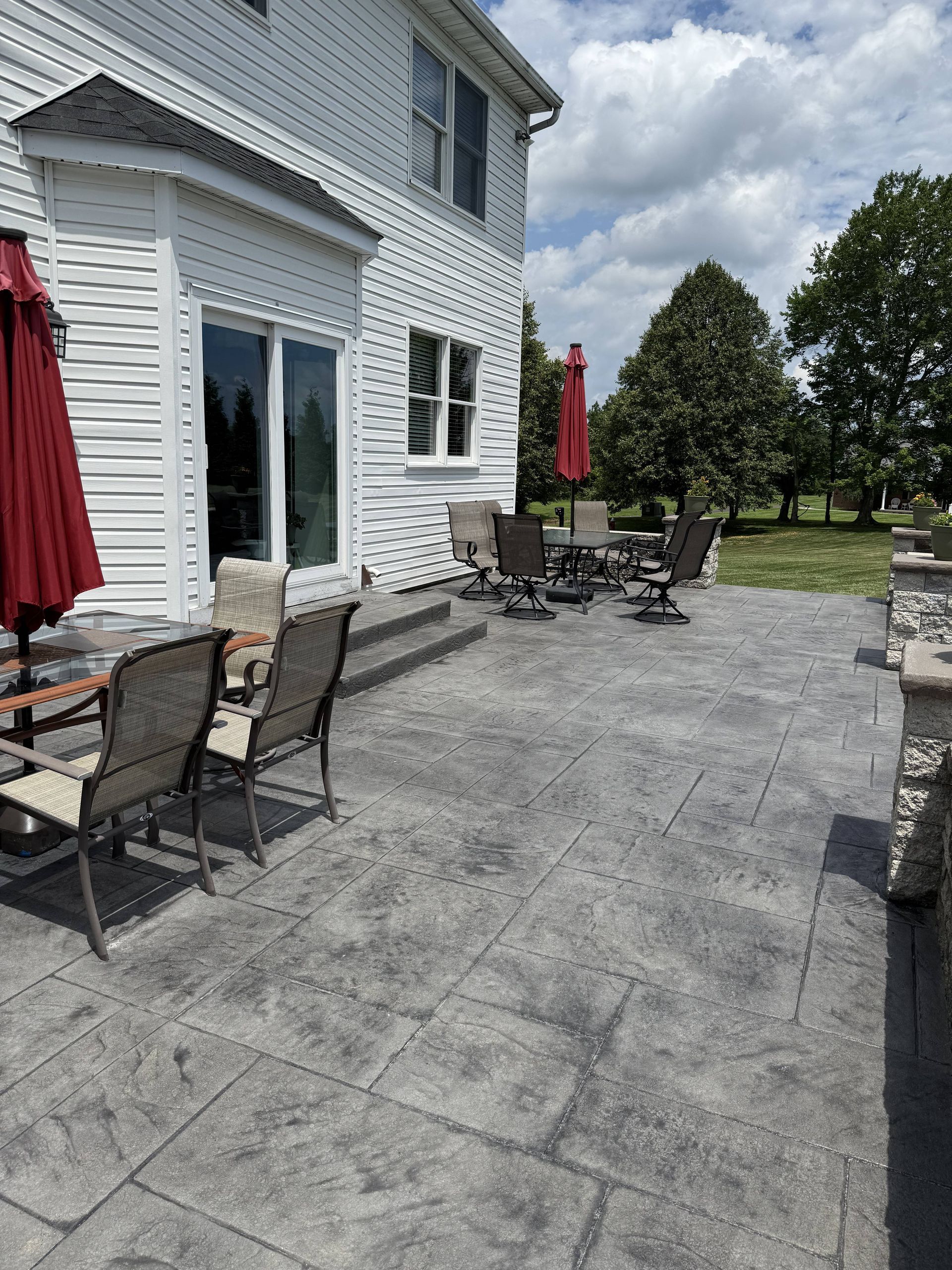 Back patio with stamped concrete, furniture, and red umbrellas next to a two-story white house on a sunny day.