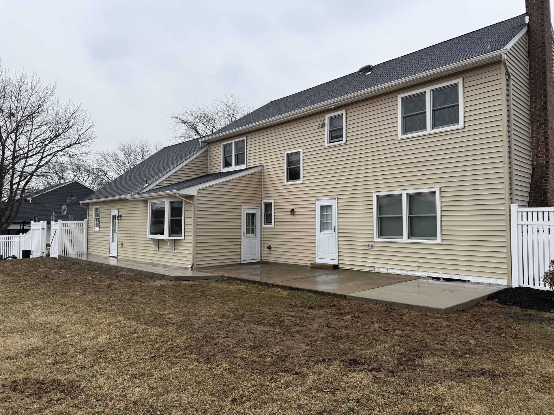 Back of a two-story beige house with a brick chimney. A concrete patio is in front of the back doors and windows.