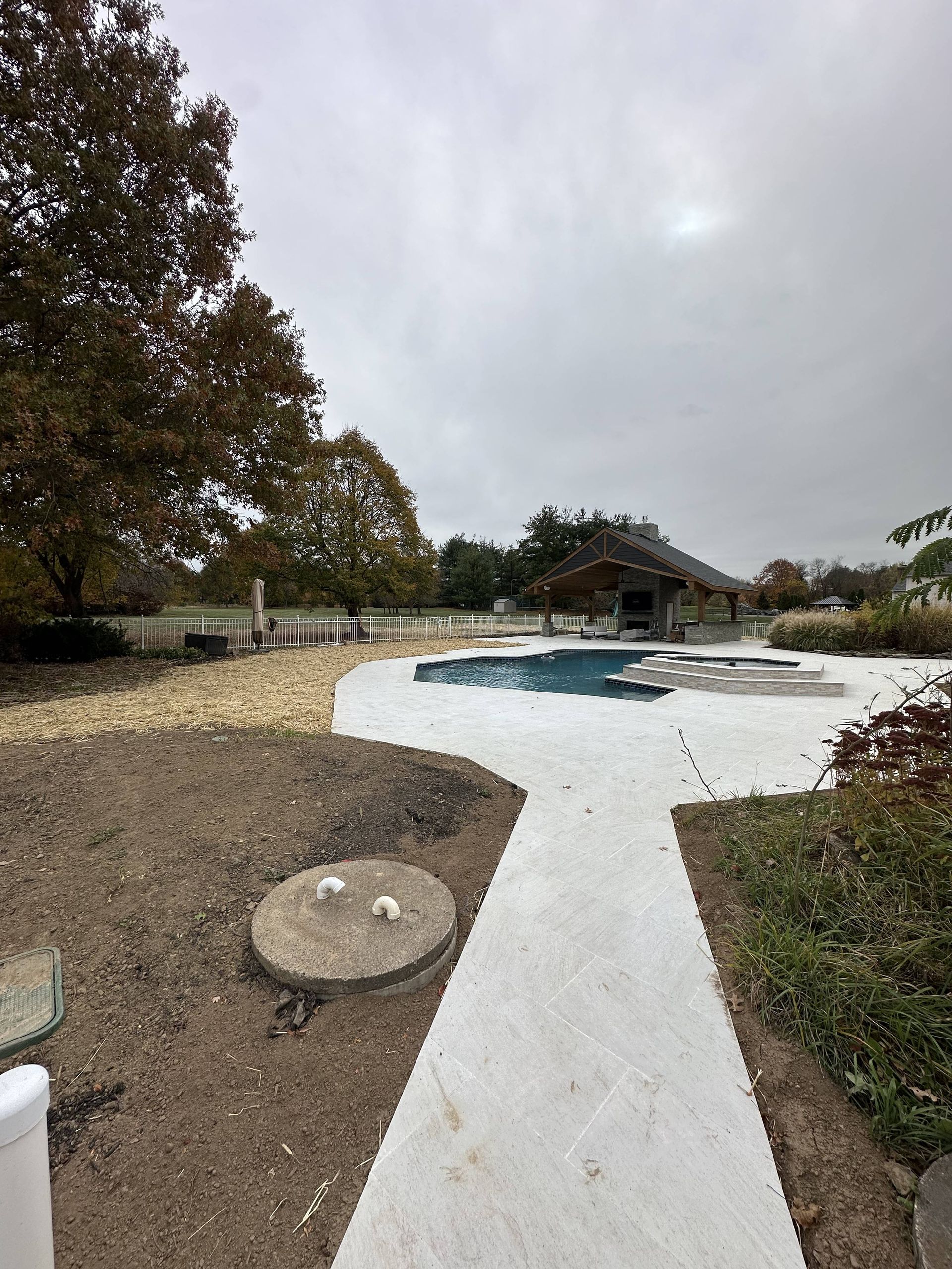 Cloudy day view of a backyard with a pool, patio, a small structure, and trees.