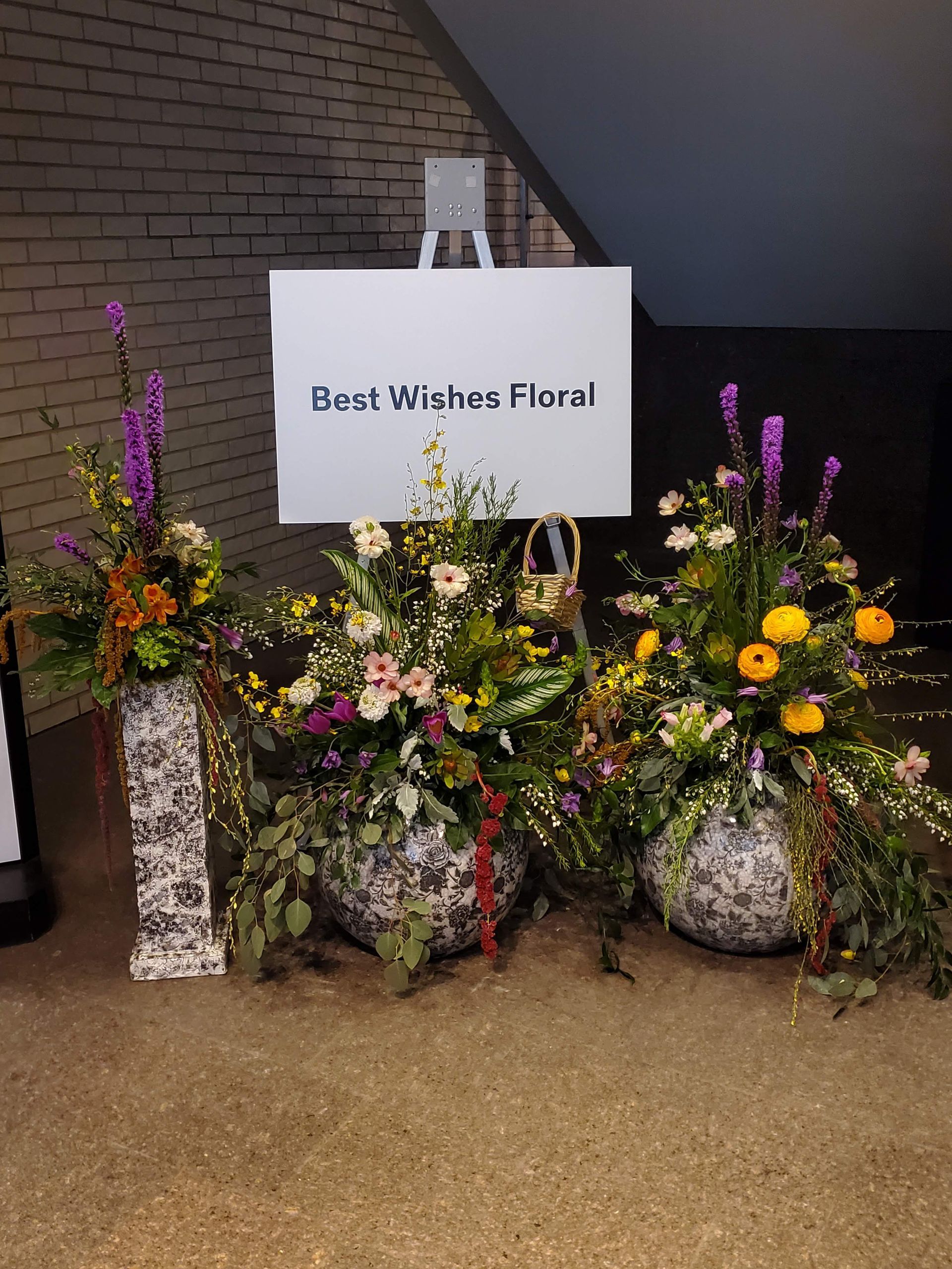 A sign that says best wishes floral is surrounded by potted flowers.