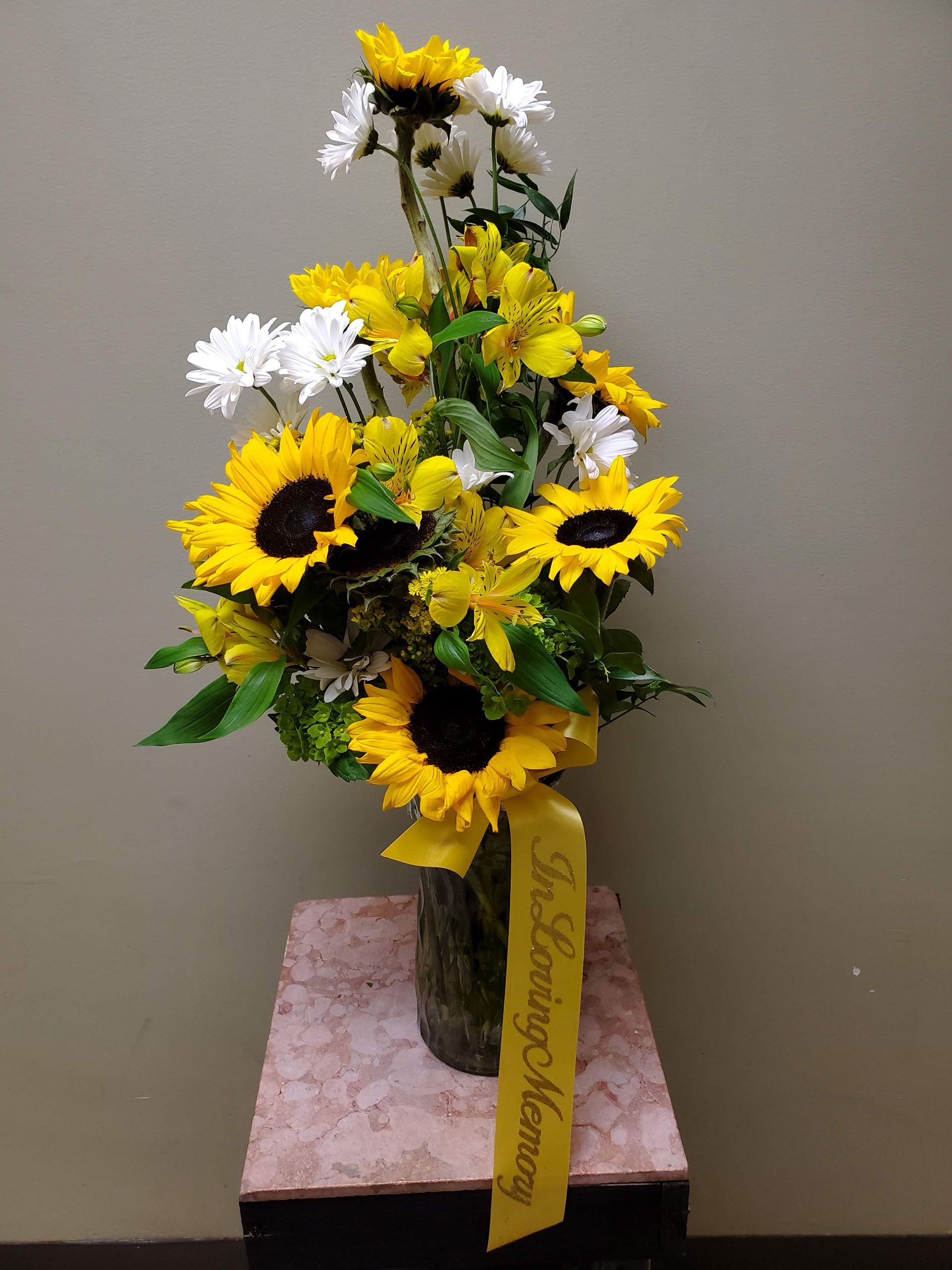A vase filled with sunflowers and daisies with a yellow ribbon that says
