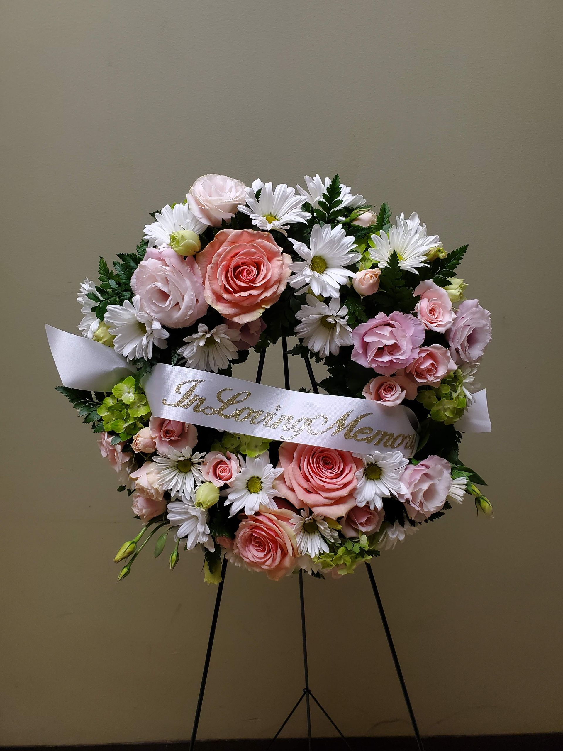 A wreath of pink and white flowers with a white ribbon on a stand.